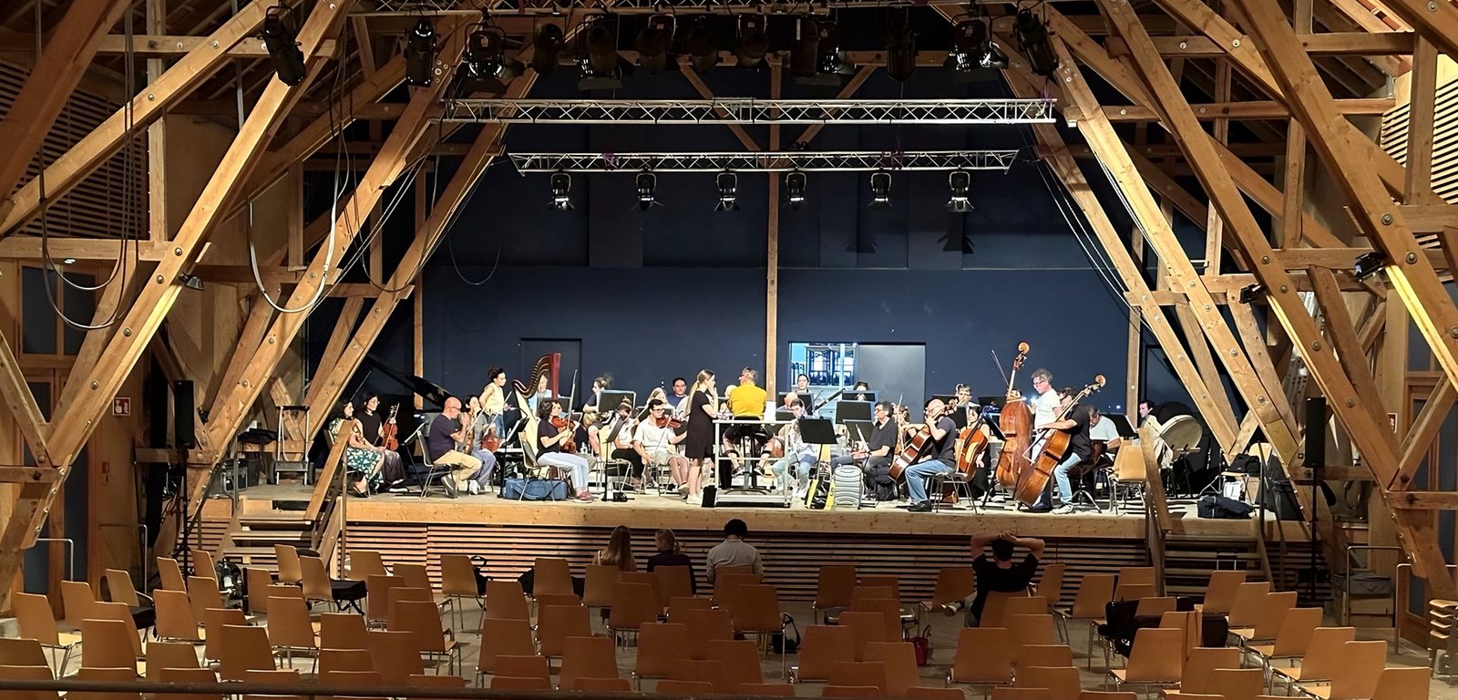 Orchester auf der Bühne eines Saals mit offenem Holzdachstuhl und Lichttechnik