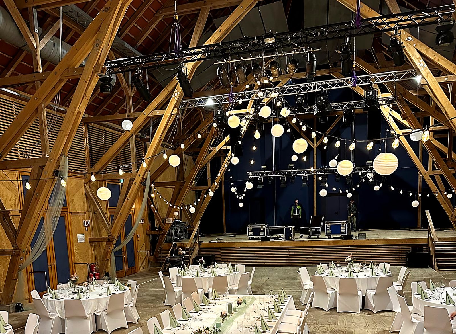 Festlich gedeckter Saal mit langer Tafel, Lampions und Lichterketten unter einem offenen Holzdachstuhl